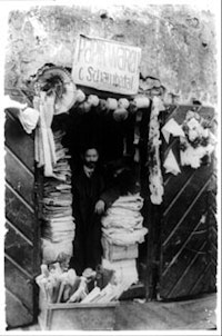 Man with papers and paper decorations in doorway under sign “Papirwaren, C. Schambedal” (papershop) in Jewish ghetto, Vilna, Russia. (1922). Courtesy of The Library of Congress.