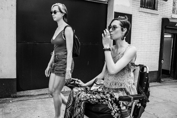 Audre Wirtanen and Laura Tuthall, 2019. Photo: Bryan Bisordi.<br><em>A black and white photo taken on a hot NYC summer day. Audre and Laura are in profile on a mission down the sidewalk. Audre, in the background, walks beside Laura rolling in their wheelchair. They both wear sunglasses, summer clothes, and short hair. They are both Hypermobile, white, and femme. Laura inhales from a medical marijuana vaporizer as they traverse together, pensive, serious, and sweaty.</em>